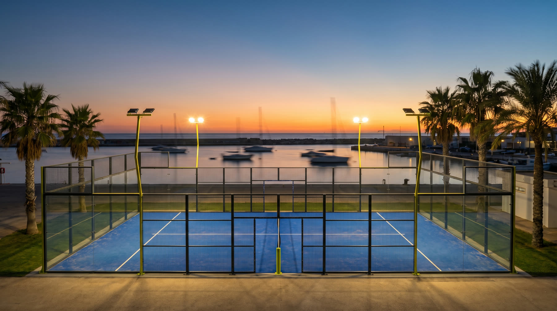 A floodlit padel court with glass walls beside a harbour at dusk, palm trees on either side.